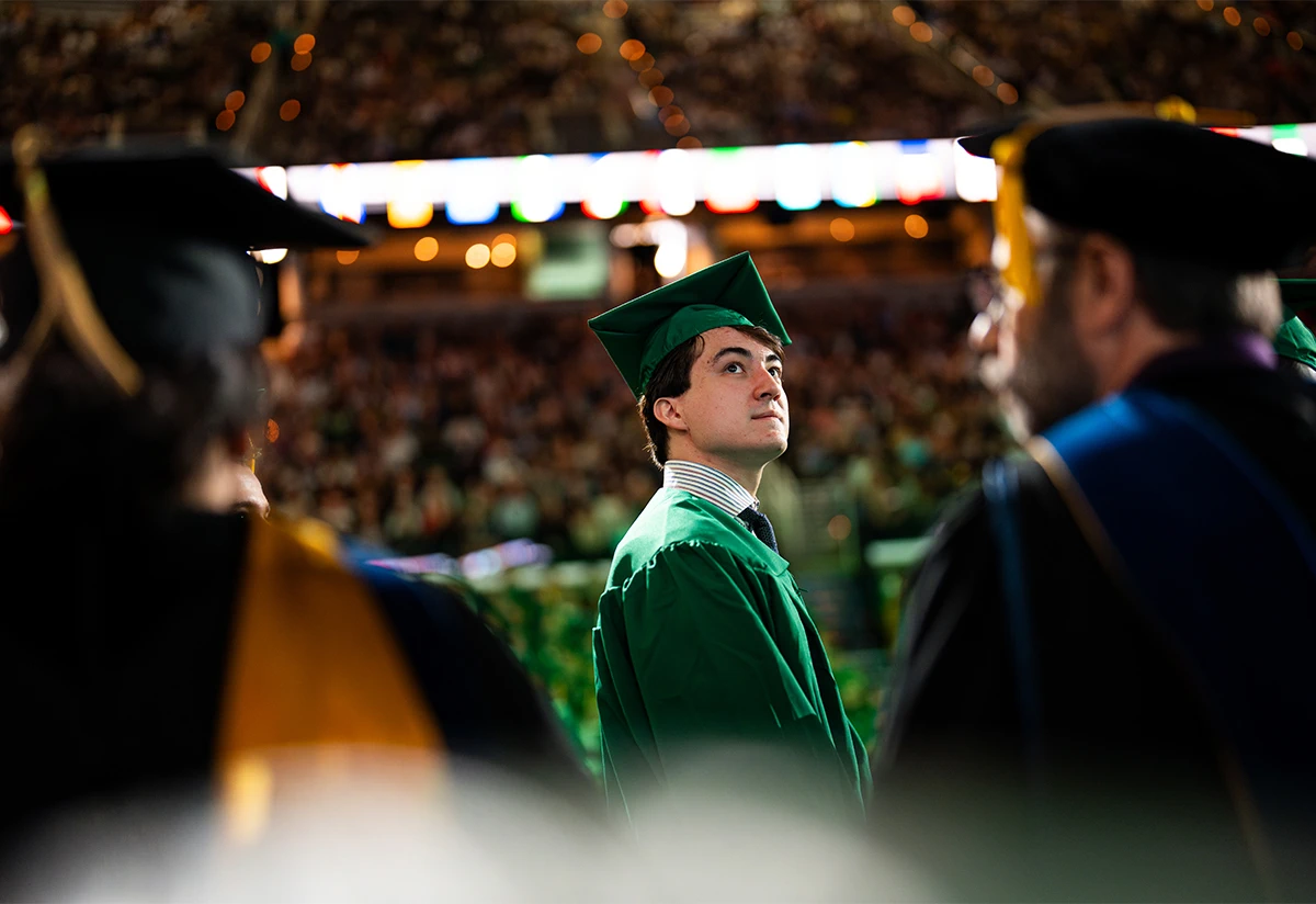a graduate is standing up while wearing a green graduation gown at the Breslin Center surrounded by others graduates